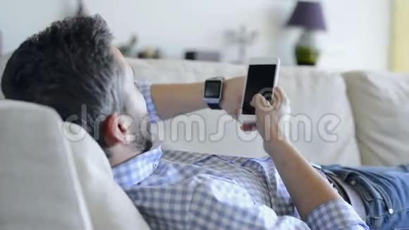 男人躺在沙发上用智能手机和智能手表视频的预览图