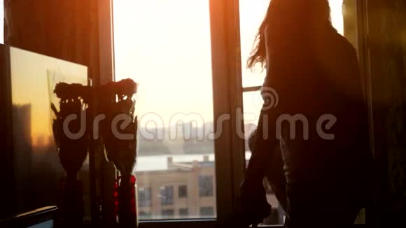 坐在窗户上的漂亮年轻的黑发女人在日落和城市的背景下使用智能手机3840x2160视频的预览图