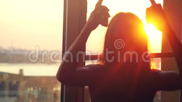 年轻的白日做梦的女人坐在窗前戴着耳机穿过太阳在令人惊叹的日落中听音乐视频的预览图