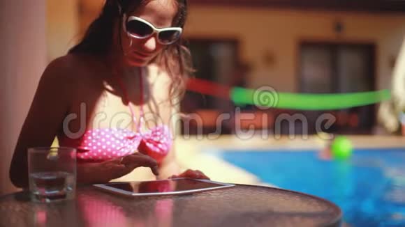 穿着太阳镜和比基尼的女孩在游泳池旁边放了一块平板电脑1920x1080视频的预览图