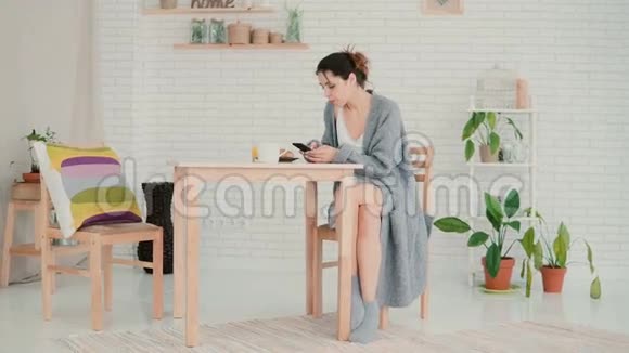 快乐的女人穿着睡衣坐在厨房里上网布鲁内特女孩使用智能手机喝咖啡视频的预览图