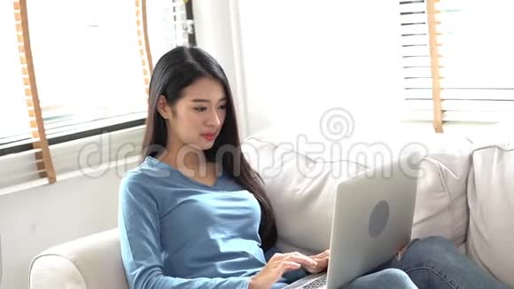 肖像美丽的亚洲年轻女子坐在沙发上在家里使用笔记本电脑休闲放松视频的预览图