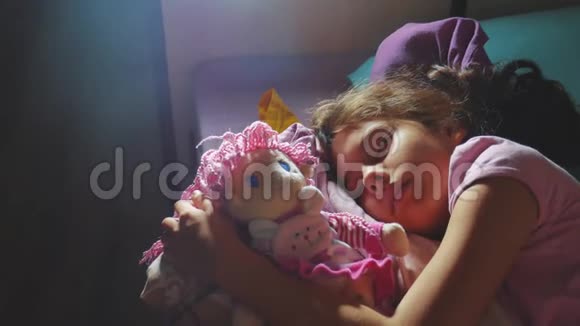 可爱的小女孩睡在沙发床上拥抱她的娃娃睡在夜间生活方式中的黑发小女孩视频的预览图