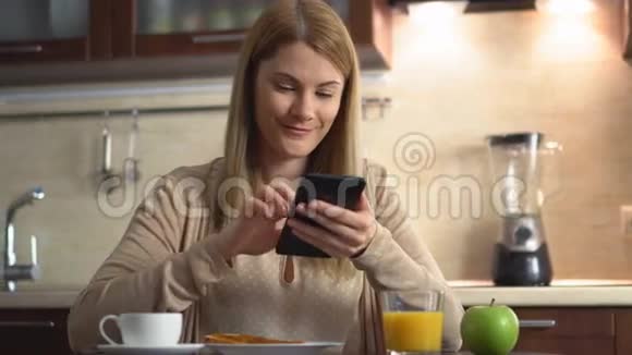 美丽的年轻迷人的女人吃早餐在厨房里浏览互联网视频的预览图