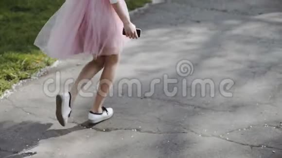 阳光明媚的一天漂亮的年轻女人穿着粉红色的裙子穿着白色的衬衫在公园里戴着智能手机三轴视频的预览图