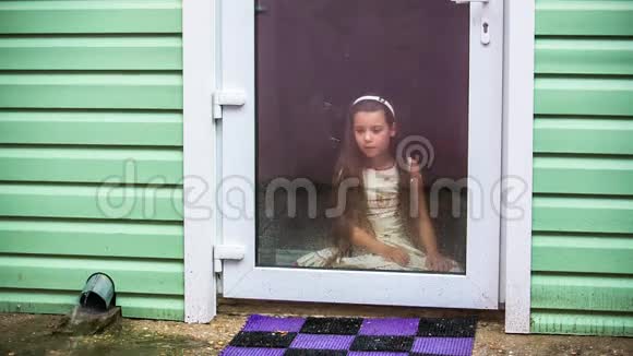 悲伤的女孩坐在玻璃门上看着雨视频的预览图