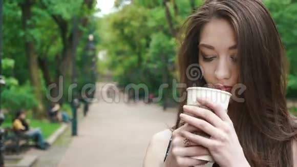 美丽的黑发女孩带着外卖咖啡走在绿色公园的街道上温暖的阳光天气户外活动视频的预览图