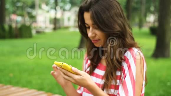 一位年轻的微笑女士她用智能手机坐在公园的长椅上视频的预览图