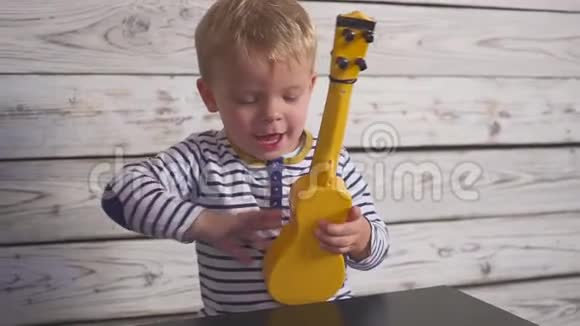 快乐的一岁男孩弹吉他或四弦琴唱歌坐在木房里视频的预览图
