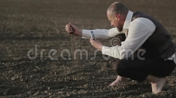 一个留胡子的人带着手机在田野里的肖像一个人光着脚在田野里给植物拍照视频的预览图