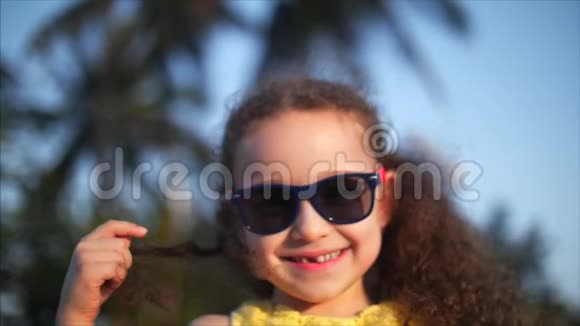 快乐的女婴穿着黄色的连衣裙和太阳镜卷发看着相机微笑着有点害羞儿童视频的预览图
