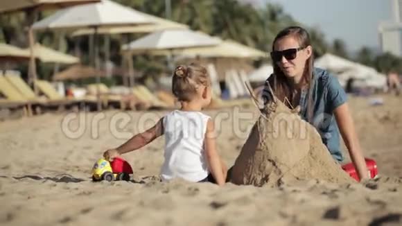 年轻的母亲穿着衣服小女儿在海滩上用沙子建造城堡视频的预览图