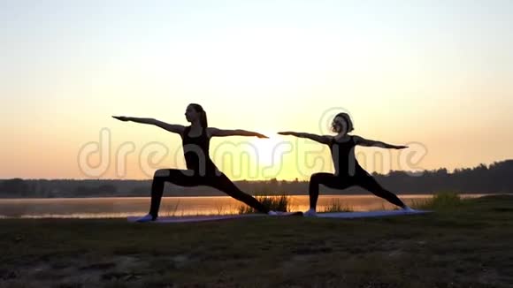 日落时两位年轻女子在风景如画的湖岸练习瑜伽视频的预览图