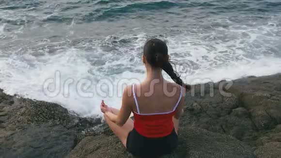 平静的女人在暴风雨的海洋前的岩石海滩上练习瑜伽健身运动视频的预览图