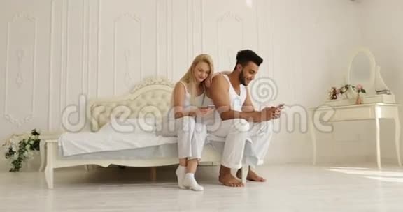 情侣使用手机坐床混搭男人女人拥抱微笑早间卧室视频的预览图