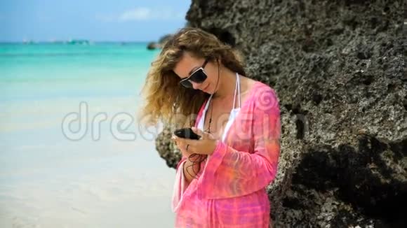 穿着鲜艳衣服的女孩在石头上的白色海滩上听音乐在海边把耳机放进耳朵的年轻女人视频的预览图
