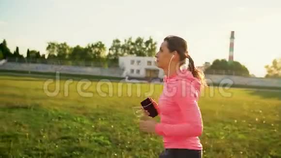 带着耳机和智能手机的女人在日落时分穿过体育场慢动作视频的预览图