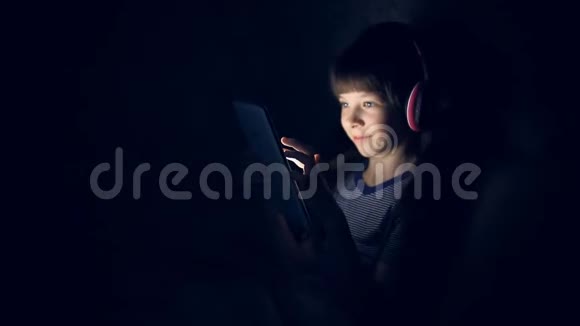 戴着无线耳机的孩子晚上在毯子下听音乐看着平板电脑视频的预览图