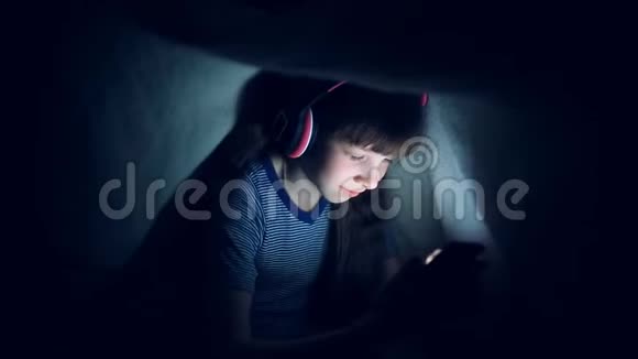 戴着无线耳机的孩子晚上在毯子下听音乐看着平板电脑视频的预览图