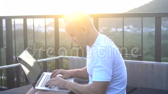 一位英俊的年轻人坐在美丽的日出的屋顶上在笔记本电脑上工作视频的预览图