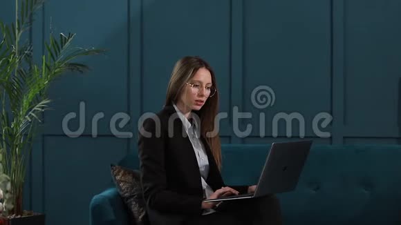 一位优雅的女商人坐在豪华蓝色办公室内部的沙发上手提电脑的肖像视频的预览图