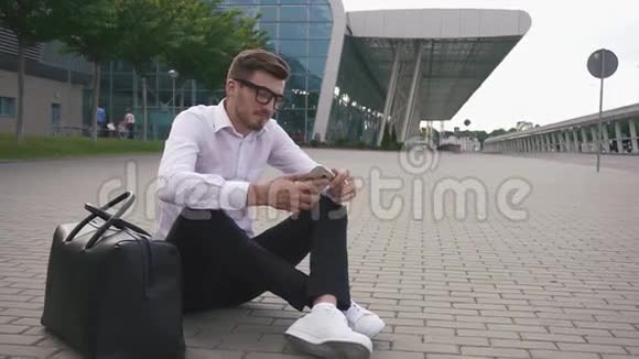 自信英俊的留着胡子的商人戴着眼镜坐在机场的地上拿着行李电话和视频的预览图