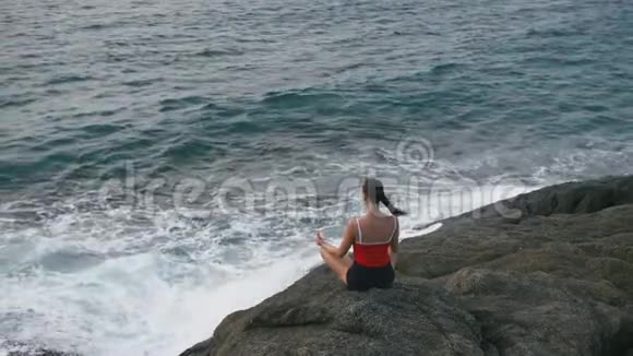 平静的女人在暴风雨的海洋前的岩石海滩上练习瑜伽健身运动视频的预览图