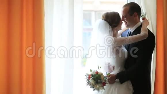 美丽的年轻夫妇迷人的新娘和可爱的新郎用橙色的窗帘在窗前拥抱和亲吻视频的预览图