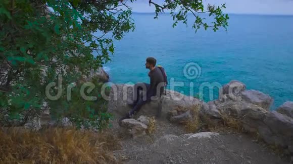 年轻的嬉皮士坐在海边的岩石山上背上背着背包的男人视频的预览图