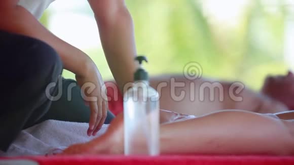 在热带海滩上一位年轻女士躺在水疗沙龙里按摩治疗师做腿油按摩1920x1080视频的预览图