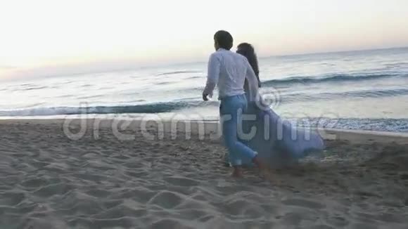 年轻浪漫的一对漂亮的女孩穿着轻盈的长裙英俊的男人在沙滩上沿着海岸嬉戏视频的预览图