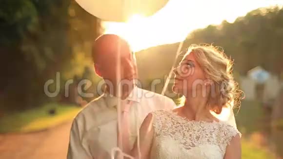 在秋天的公园里一对年轻的新婚夫妇带着气球做着滑稽的脸在夕阳的背景下笑着视频的预览图