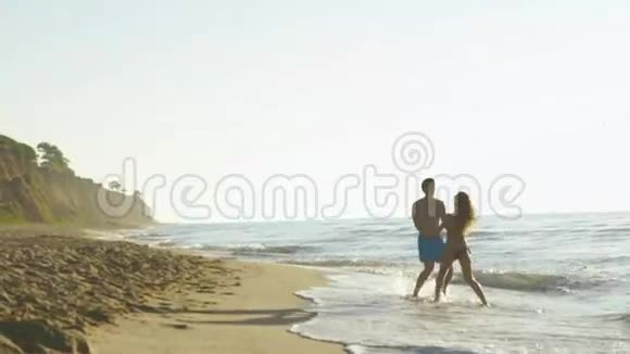 快乐的年轻夫妇在海边玩海浪在沙滩上英俊的男人和漂亮的女孩视频的预览图