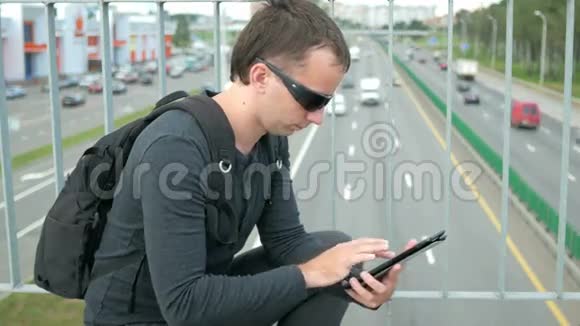 街上有数字平板电脑的现代年轻人的户外肖像戴眼镜的人背着背包坐在上面视频的预览图