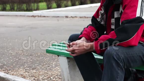 一个孤独的人坐在公园的长凳上在智能手机上打字视频的预览图