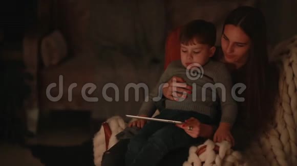 美丽的母亲坐在椅子上他的家和儿子搭在腿上在触摸屏上看动画片视频的预览图