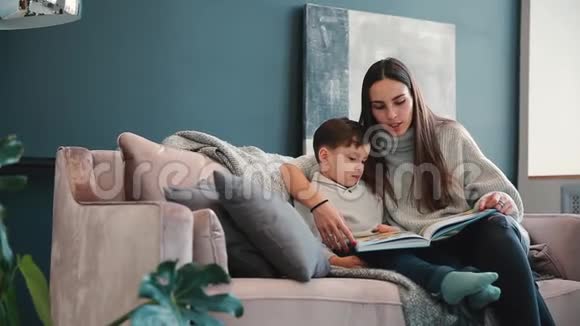 漂亮的年轻母亲坐在你家的沙发上客厅里读着儿子的耳朵里的故事学着读视频的预览图