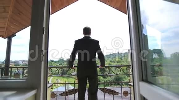 穿着黑色西装的时髦新郎站在阳台上靠在栏杆上看着背景上的绿色森林视频的预览图