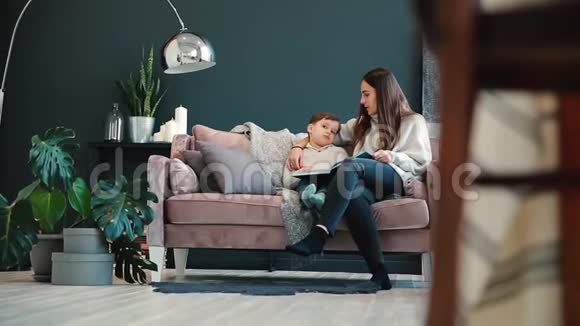 漂亮的年轻母亲坐在你家的沙发上客厅里读着儿子的耳朵里的故事学着读视频的预览图