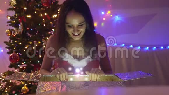 年轻快乐的美丽女子坐在神奇的圣诞礼物和圣诞树前慢慢地看着闪闪发光的灯光视频的预览图