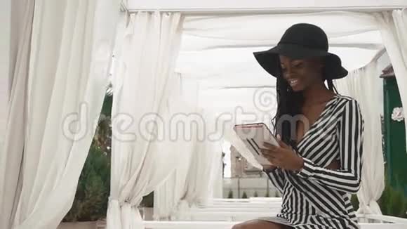 穿着条纹鸡尾酒连衣裙和戴着黑色帽子的迷人非裔美国女士在豪华酒店使用平板电脑视频的预览图