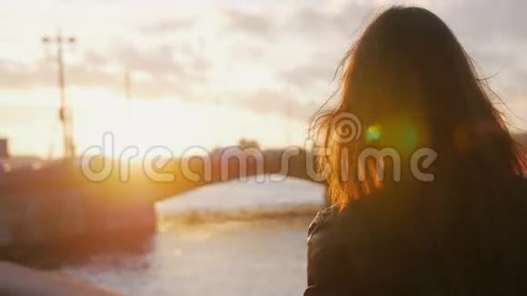 站在河堤上的年轻女子在夕阳的明亮光线下拍摄了这座桥的智能手机照片4K视频的预览图
