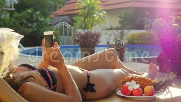 暑假戴帽子的年轻女子躺在日光浴床上在游泳池旁使用智能手机带着美丽的风景视频的预览图