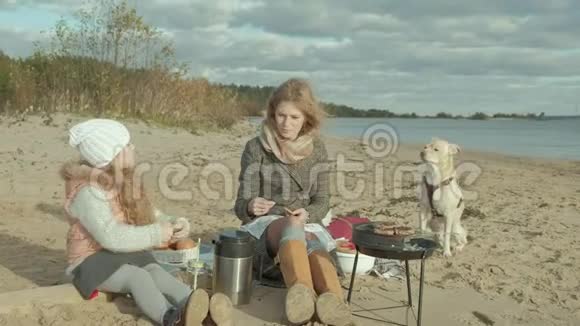 年轻的女人穿着外套一个卷发的女孩母亲带着一个女儿坐在河边的海滩上大海曾经视频的预览图
