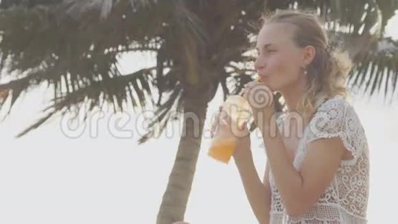 年轻女子坐在沙滩上晒太阳用吸管喝鸡尾酒视频的预览图