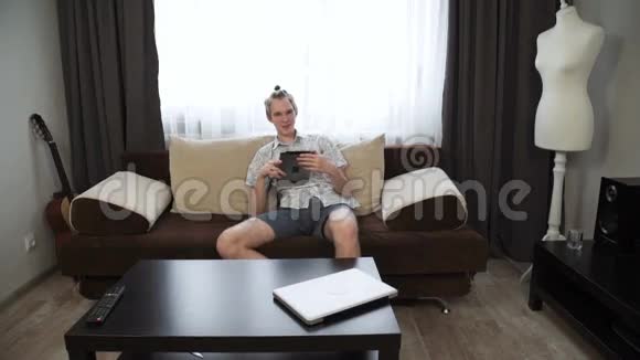 年轻人穿着短裤和衬衫头上有一条尾巴坐在沙发上上面有数字平板电脑视频的预览图