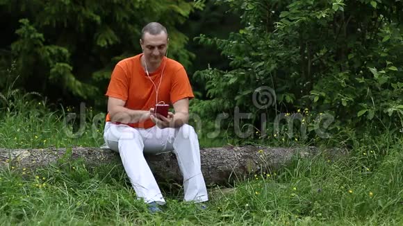 男人坐在原木上拿着电话耳机听音乐微笑视频的预览图