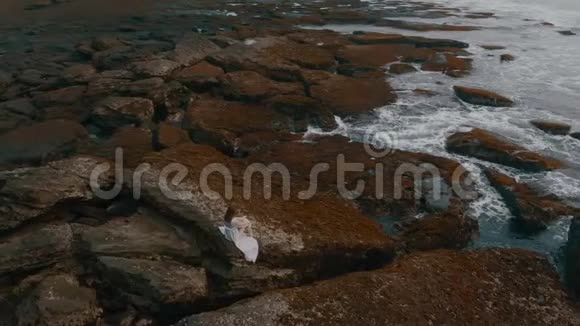 日落时分美丽的年轻女性身着长裙在巴厘岛海边的岩石上摆姿势飞机视频4K视频的预览图