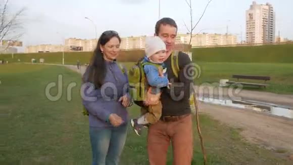 春天活跃的家庭带着婴儿在公园里散步绿草和夕阳绿色背包视频的预览图