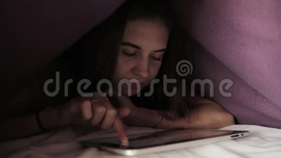 年轻女孩躲在羽绒被下在黑暗中使用PC平板设备青少年晚上在数字平板电脑上玩视频的预览图